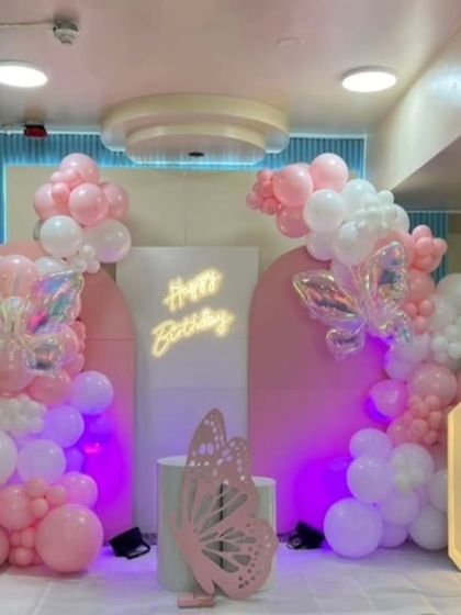 A pink and white butterfly-themed setup for a 6th birthday. The decor includes a "Happy Birthday" neon sign, iridescent butterfly balloons, and a large marquee number "6".