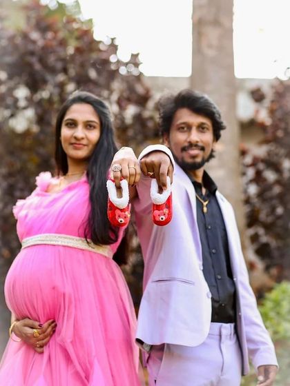 A sweet couple's maternity photo announcing their baby's arrival with tiny shoes. The pink gown and the partner's light-colored suit make a perfect pair.
