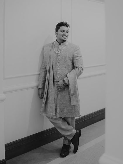 A classic black and white portrait of the groom leaning against a wall. His relaxed pose and confident smile create a timeless and effortlessly cool image.