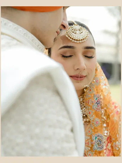 A moment of pure serenity. This intimate close-up focuses on the bride's peaceful expression as the groom kisses her forehead. Her look is styled with a statement maang tikka and soft, natural makeup.