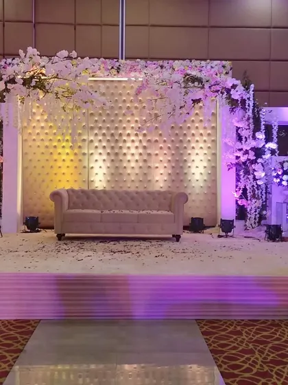 A modern pastel stage setup inside a banquet hall. The backdrop features a textured white panel, accented with an arch of cherry blossom flowers and soft purple lighting.