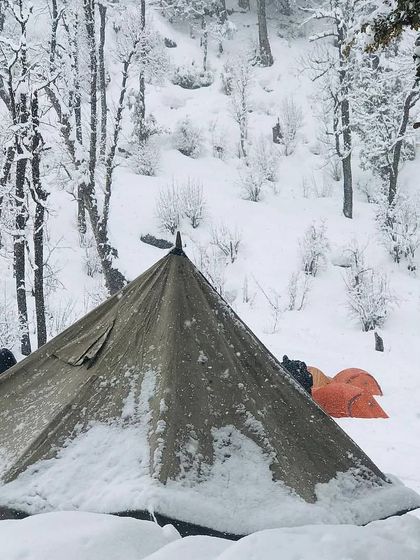 A classic canvas tent covered in a layer of fresh snow, with other dome tents in the background. This highlights the different types of gear used for a comfortable winter camping experience.