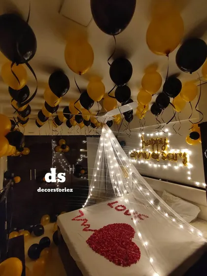Surprise your loved one with a romantic bedroom transformation. This anniversary setup features a fairy-light canopy over the bed, a heart made of rose petals, a 'Happy Anniversary' foil balloon banner, and dozens of balloons on the ceiling and floor.