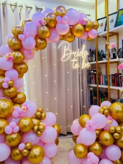 I design stylish and fun backdrops for bridal showers. This setup uses a modern gold metal arch decorated with an arrangement of pink and metallic gold balloons, complemented by a "Bride to be" neon sign and delicate fairy lights to create a warm, celebratory glow.