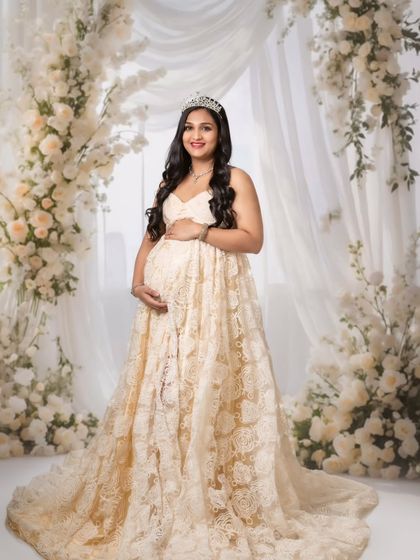 A classic portrait of a mom-to-be in a beautiful lace gown, wearing a tiara against a wall of white flowers for a truly royal feel.
