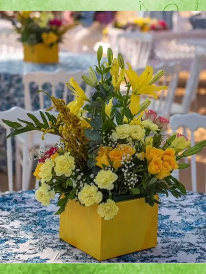 A close-up of a floral centerpiece from a traditional wedding. The bright yellow lilies and carnations in a matching yellow vase add a pop of color to the guest tables.