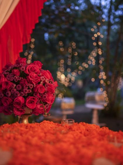 A beautiful evening shot of the Mehendi decor, with a bouquet of red roses in the foreground and the soft, blurred bokeh of fairy lights in the trees behind.