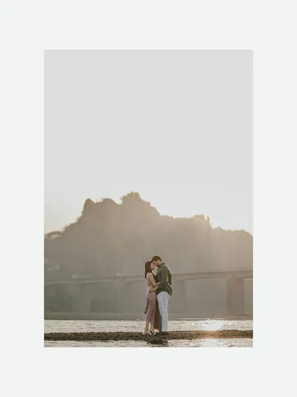 A wide, atmospheric shot of the couple sharing an intimate moment on a sandbar. The golden sunset and distant bridge create a beautiful, cinematic backdrop for their beach pre-wedding session.