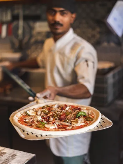 A chef presents a fully topped pizza on a peel, ready for the intense heat of the wood-fired oven. This is the last moment before it transforms into a crispy, bubbly masterpiece.