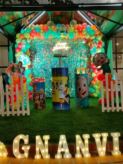 A Cocomelon party at night, with a bright sequin wall and neon sign creating a vibrant glow. The setup is on artificial grass with a white picket fence.