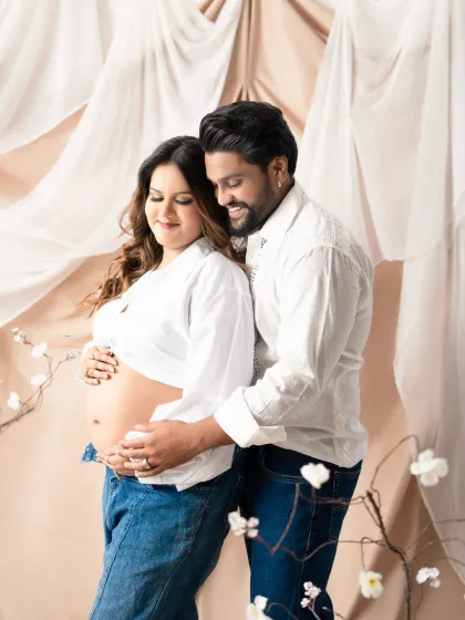 A loving embrace during a relaxed, casual maternity session. The couple's coordinated white shirts and jeans create a clean and timeless look against the soft, draped backdrop.