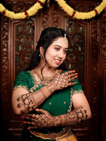 A smiling bride showcasing her full bridal mehendi. The combination of her green saree and the red henna stain is a classic and beautiful look.