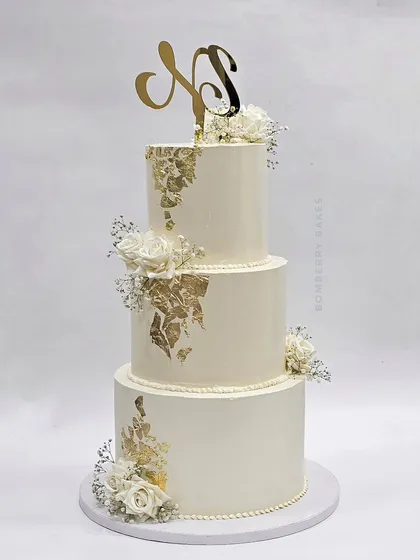 A classic three-tier white and gold leaf wedding cake, adorned with delicate white roses and baby's breath. The custom monogram topper adds a personal touch.