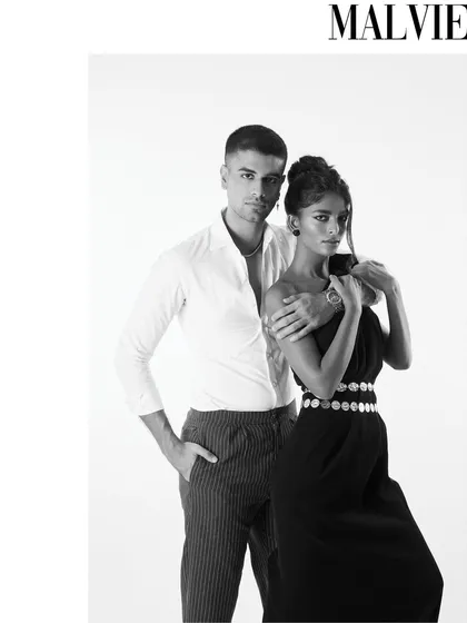 A full-length black and white portrait showing the couple's modern styling. The contrast between his simple shirt and her elegant dress with a coin belt creates a balanced and fashionable composition for this editorial.
