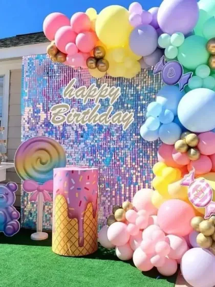 A candy-themed birthday party with a rainbow sequin wall. This outdoor setup features a pastel balloon arch and large lollipop and teddy bear props.