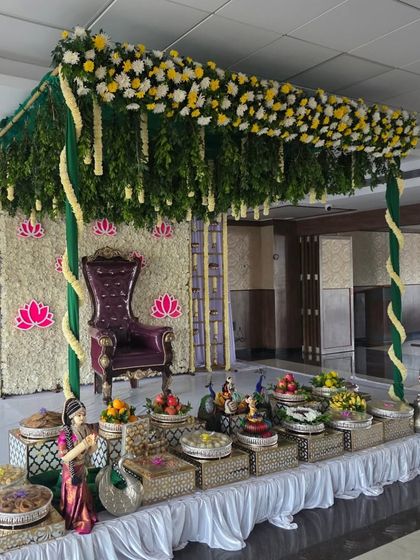 A half-saree ceremony setup in a hall, featuring a floral canopy, a jasmine wall with lotus motifs, and an extensive offering table.