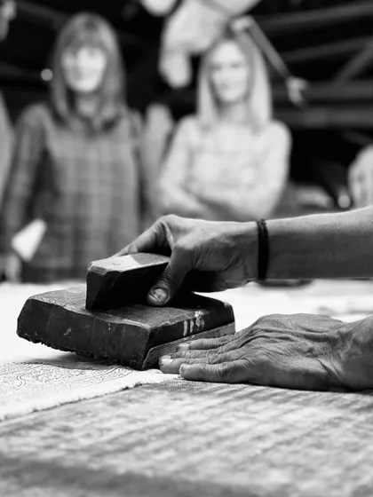 A black and white photograph capturing the essence of our craft. An artisan's hands guide the block, a skill honed over many years, as visitors watch in the background during a studio tour.