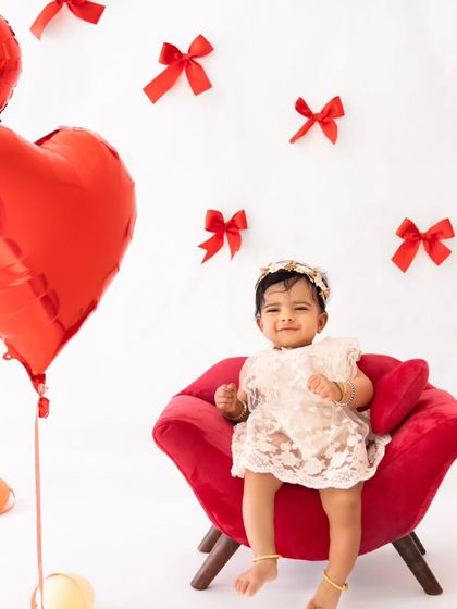 A sweet smile from the baby girl, surrounded by red heart balloons in her adorable Valentine's Day setup.