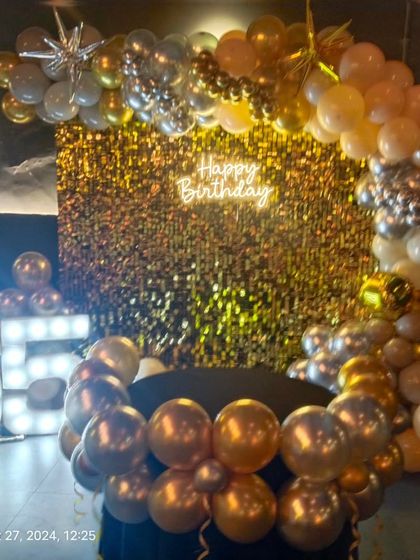 A glamorous 65th birthday celebration with a gold shimmer wall, a balloon garland in gold and silver, and large light-up marquee numbers.