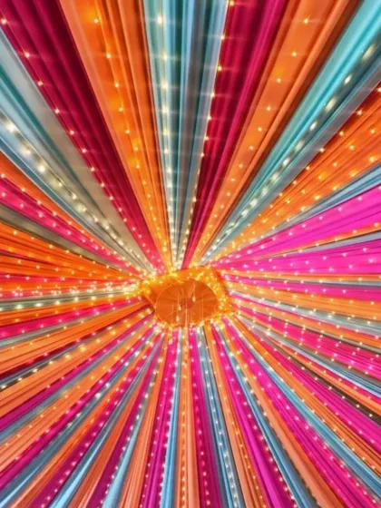 A close-up view of a vibrant ceiling canopy. The combination of bright orange, pink, and blue drapes with strings of fairy lights creates a warm and celebratory glow for any evening event.