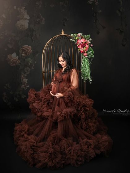 A quiet moment of anticipation. Seated by the golden birdcage, the mom-to-be looks down at her bump with love, wrapped in a voluminous brown gown.