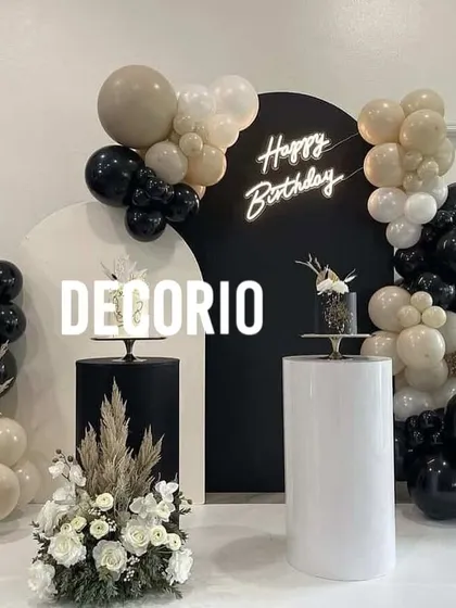 A modern and sophisticated birthday setup featuring a bold black circular backdrop with a custom 'Happy Birthday' neon sign. The scene is balanced with organic balloon garlands in black, white, and neutral tones, and includes elegant white and black pedestals for cakes.