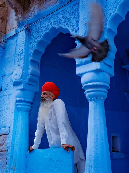 A man in a traditional red turban stands in a classic blue archway in Jodhpur as a pigeon takes flight. This shot captures the perfect contrast of color and a fleeting moment of life in Rajasthan's Blue City.