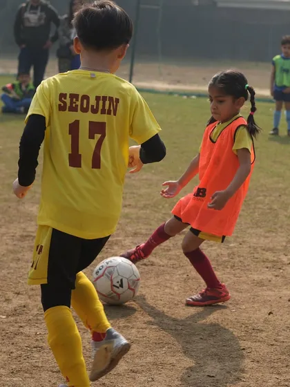 A young girl in an orange bib challenges a boy for the ball. We create many firsts for our players, including their first taste of healthy competition.