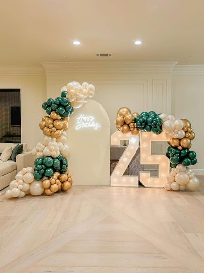 A chic and modern setup for a 25th birthday. This decoration features organic balloon arrangements in emerald green, gold, and cream, an arched backdrop with a neon sign, and large marquee numbers. It's a stylish way to celebrate a quarter-century.