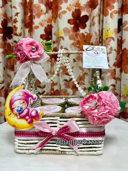 A lovely baby girl hamper in a white wicker basket, decorated with pink flowers and a 'baby on the moon' cutout.