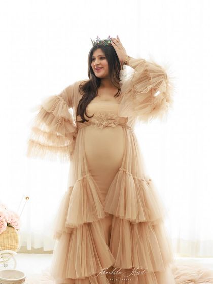 A joyful, regal portrait of a mother-to-be adjusting her crown, smiling in a beautiful nude-toned ruffled gown.