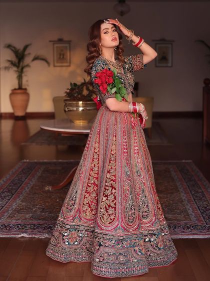 A full-length shot of a bride in a multi-colored lehenga with a green blouse, holding red roses. The combination of colors is striking and unique.