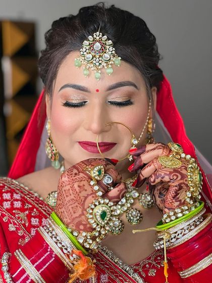 A stunning bride in a classic red lehenga, her hands framing her face to show off the intricate henna and jewellery. The makeup is bold and glamorous.
