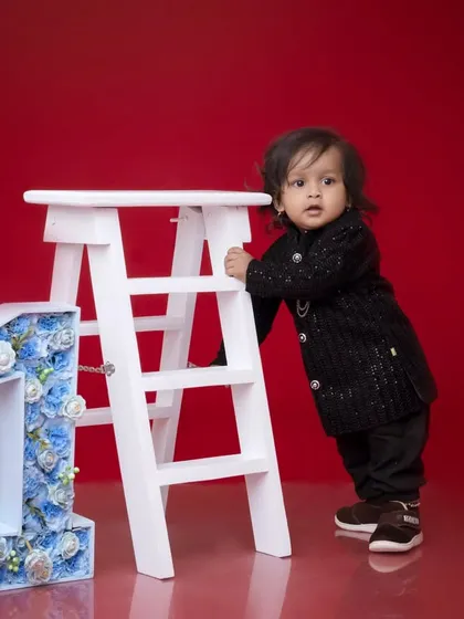 A stylish first birthday portrait against a bold red background. The baby boy leans against a white ladder next to a floral number '1', creating a modern and striking image.