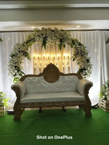 A clean and classic reception stage setup. The white floral arch against a white drape, paired with a simple white sofa, creates a timeless and elegant look.