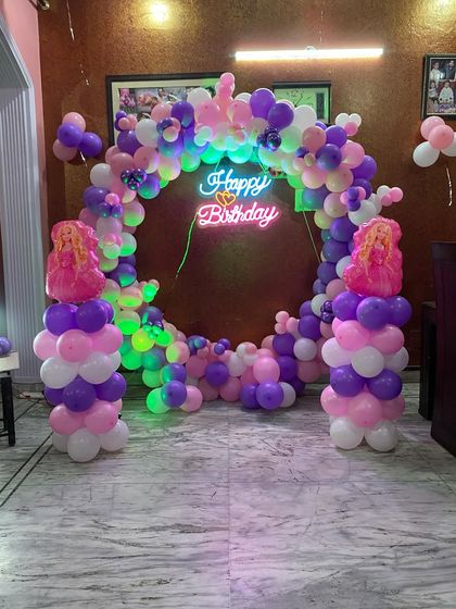 A princess-themed birthday party decoration with a circular balloon arch in pink, purple, and white. The arch is lit with a neon sign and features princess figures on the side pillars.