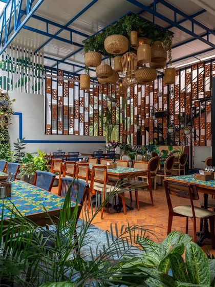 This tropical-themed restaurant space features an earthy color palette with pops of blue and plenty of greenery. The custom wooden screen and hanging cane lights add warmth and texture, creating a relaxed and welcoming atmosphere.