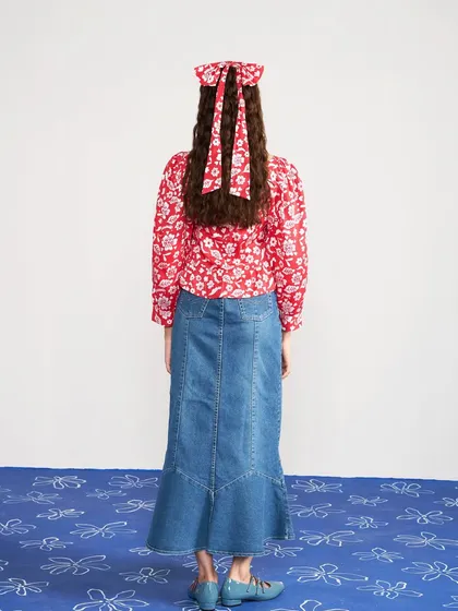 A back view of a look for Joskai Studio, pairing a red floral top with a denim maxi skirt. This shot highlights the bow detail in the hair and the silhouette of the skirt.
