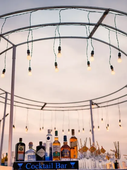 A stylish circular cocktail bar set up for a sundowner event. Edison bulbs hang from the frame, illuminating the bar and creating a warm, inviting space for guests to mingle as the sun sets over the ocean.