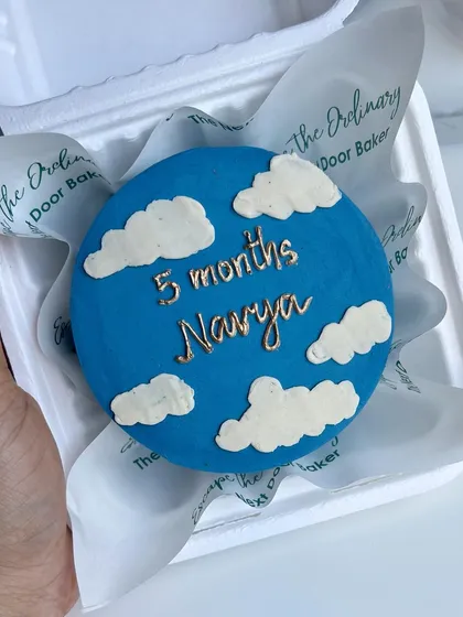 A 5-month milestone cake with a beautiful blue sky and fluffy white clouds. The name is piped in elegant gold lettering.