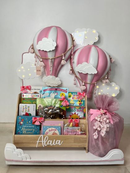 A beautiful gift set for baby Alaia, featuring a personalized bookshelf, pink hot air balloon lamps, and a large, elegantly wrapped present.