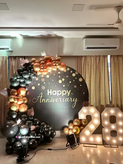 A stylish 28th-anniversary backdrop with a dark, sparkling panel. The design is accented with a cascade of black, copper, and gold balloons and large light-up marquee numbers.