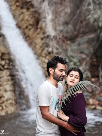 An intimate and artistic portrait by a waterfall. The use of a fern leaf adds a soft, natural element to the foreground, drawing focus to the couple's tender moment.