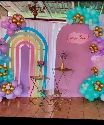 A boho-rainbow themed decoration perfect for a baby shower. This setup includes nested rainbow arch cutouts, a personalized backdrop, and balloon arrangements in a soft pastel palette of purple, teal, and gold.
