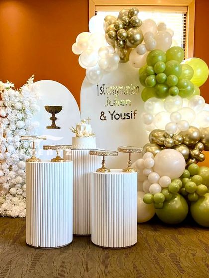 A modern First Communion decoration for a boy, featuring an olive green, white, and gold balloon arch. The setup includes a cake display on textured white plinths against a clean, arched backdrop.