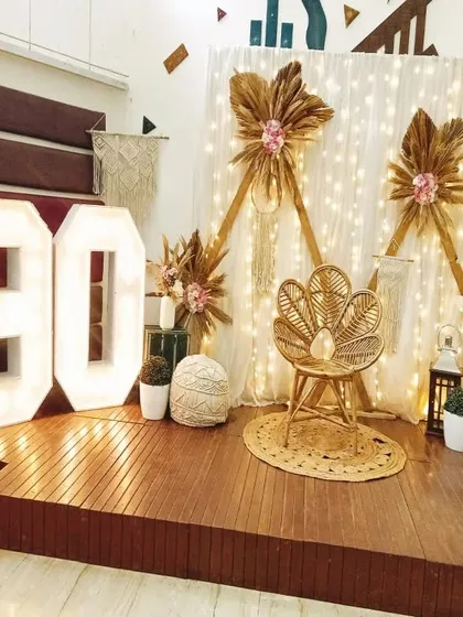 A rustic and chic 90th birthday setup on a wooden stage, featuring a peacock chair, macrame details, dried floral arrangements, and large marquee numbers.