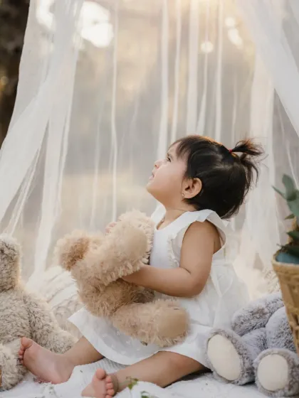 A toddler sits with her teddy bear, looking up in wonder. The soft, natural light creates a beautiful, gentle mood.