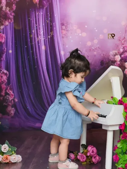 A little girl in a denim dress plays a miniature white piano in a magical, floral setting. The purple curtains and flower-draped piano create a dreamy and artistic scene.