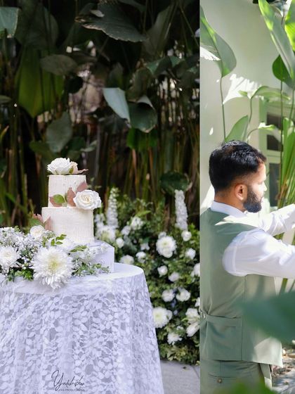 A beautiful three-tiered wedding cake adorned with fresh white roses, set on a lace-covered table. The surrounding white floral arrangements create a cohesive and elegant dessert station.