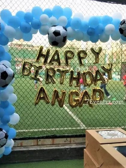 A football-themed birthday decoration set up against the net of a football pitch. The blue and white balloon arch is decorated with football foils.